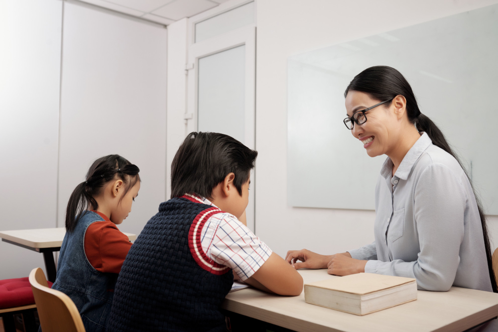 two-asian-kids-sitting-classroom-smiling-teacher-glasses-talking-boy